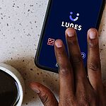 Eine Hand, die am Smartphone in der Lunes-App tippt. Daneben steht eine Tasse Kaffee. 